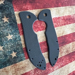 May include: Two black carbon fiber knife scales with a textured weave pattern. Each scale has a hole at the top and several smaller holes along the edges. The background is an American flag.