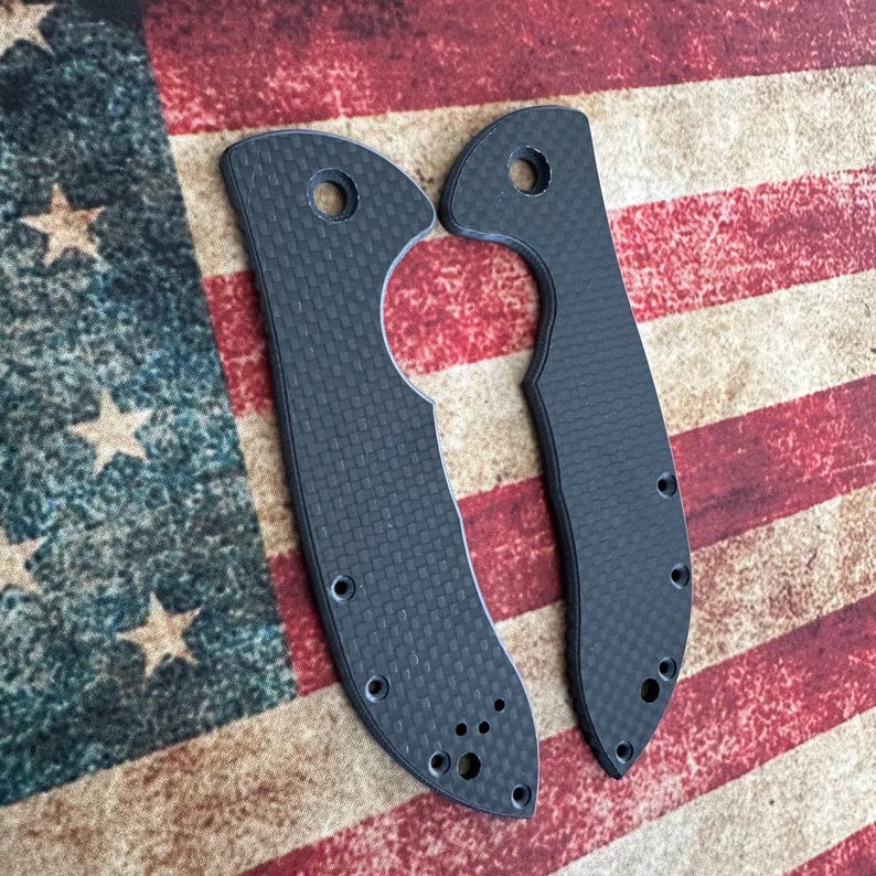 May include: Two black carbon fiber knife scales with a textured weave pattern. Each scale has a hole at the top and several smaller holes along the edges. The background is an American flag.