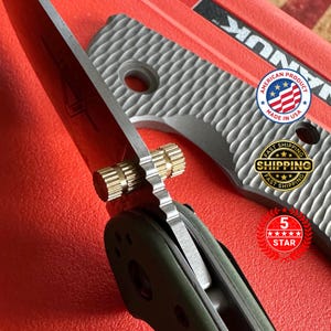 May include: Close-up of a partially open folding knife with a silver blade and a brass pivot. The handle is olive green and the textured grip is gray. The knife is set against a red background, with an "American Product Made in USA" sticker.