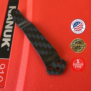 Op de afbeelding: Een zwarte koolstofvezel clip met een textuurpatroon. De clip heeft drie gaten aan de basis en is geplaatst tegen een rode achtergrond. De afbeelding bevat ook een "Made in USA" sticker.