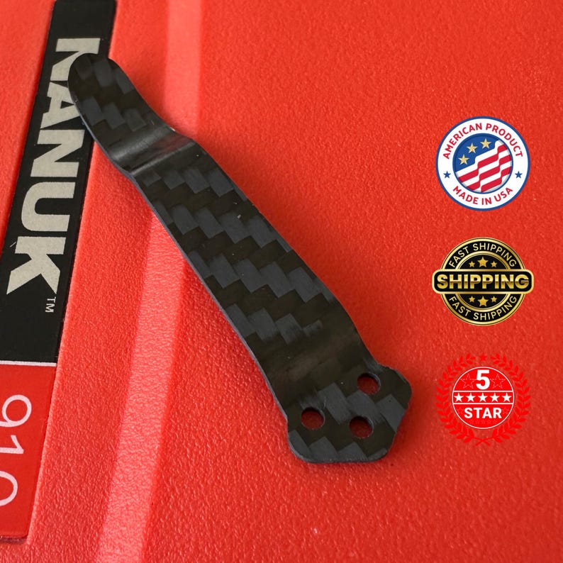 Op de afbeelding: Een zwarte koolstofvezel clip met een textuurpatroon. De clip heeft drie gaten aan de basis en is geplaatst tegen een rode achtergrond. De afbeelding bevat ook een "Made in USA" sticker.