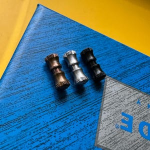 May include: Three metal fidget spinners in bronze, silver, and black. Each spinner has a cylindrical body with textured detailing on the ends. The spinners are arranged on a blue surface with a yellow background.