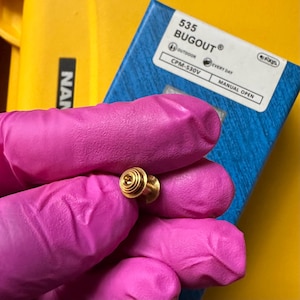 May include: A small gold-colored metal thumb stud for a folding knife, featuring a circular grooved top and a cylindrical base. In the background, a blue box displays the text 535 BUGOUT, CPM-S30V, MANUAL OPEN, and AXIS.