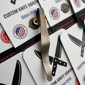 May include: A silver-colored knife clip with a brass-colored rivet is displayed against a backdrop of business cards. The cards feature the text "CUSTOM KNIFE HARDWARE Since 2018" and "AMERICAN PRODUCT MADE IN USA" logos.