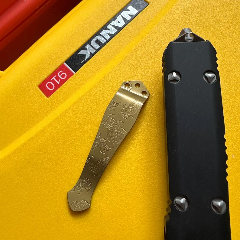May include: A gold-colored metal pocket clip featuring an engraved koi fish pattern, positioned next to a black handle of a folding knife on a yellow Nanuk 910 case. The clip has three small circular mounting holes at the top end.