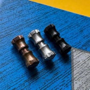 May include: Three metal fidget spinners in bronze, silver, and black. Each spinner has a textured, decorative design on the ends and a cylindrical body. The spinners are displayed on a blue surface with a yellow corner.