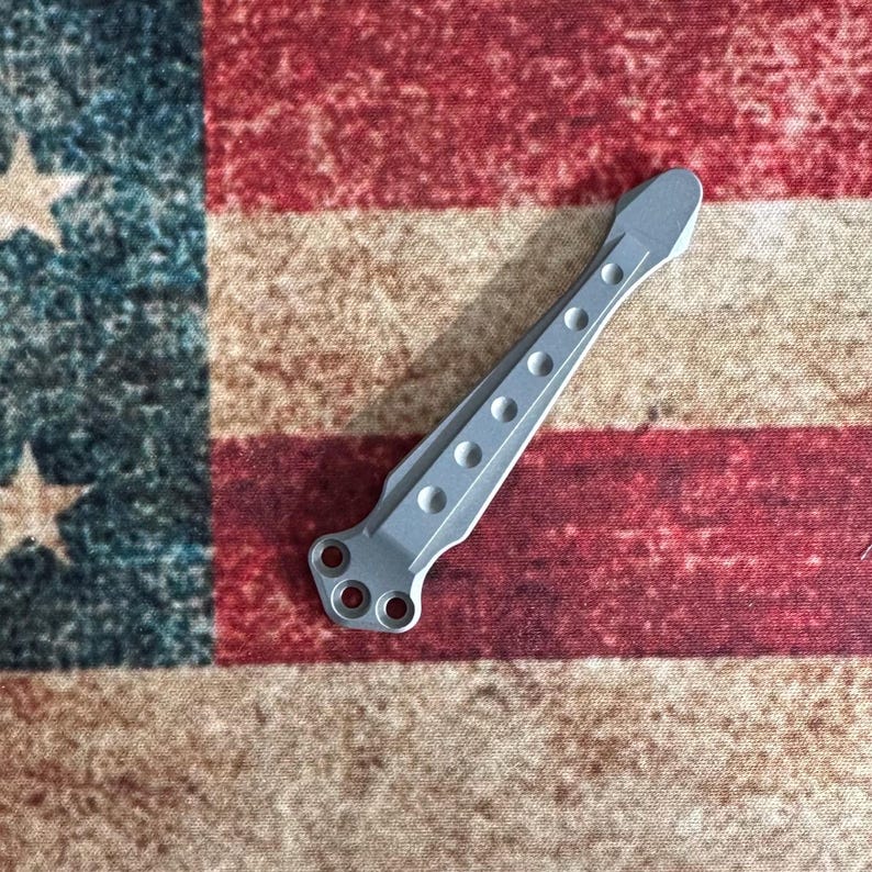 Pu&ograve; includere: Un componente metallico color argento con una serie di fori circolari e tre fori pi&ugrave; grandi a un'estremit&agrave;. Il componente &egrave; posto su uno sfondo della bandiera americana, con le sue strisce rosse e bianche e il suo campo blu stellato.