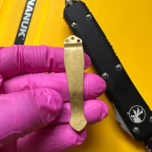 May include: A gold-colored metal pocket clip featuring an engraved koi fish pattern. The clip has two small circular holes at the top for attachment and a tapered shape. It is positioned next to a black handle of a folding knife.
