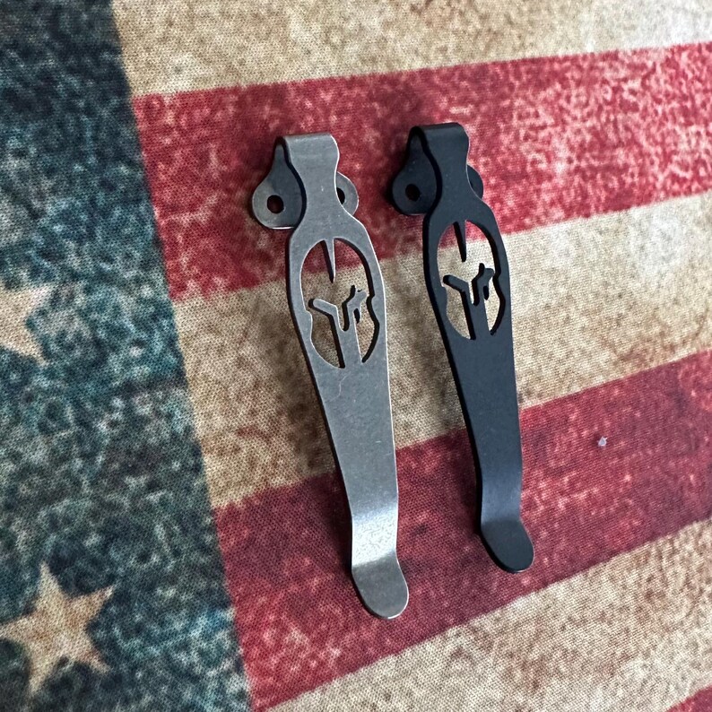 May include: Two metal pocket clips, one silver and one black, each featuring a Spartan helmet cutout design. The clips have a curved shape with a small hole at the top for attachment. The background is a fabric with a red, white, and blue pattern.