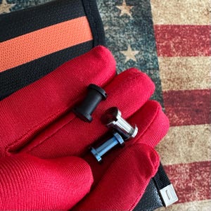 May include: Three metal plugs in black, silver, and blue are displayed on a red surface. A black wallet with an orange stripe is visible in the background, along with a faded American flag.