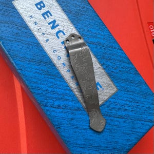 Op de afbeelding: Een zilverkleurige zakmesclip met een gegraveerd visontwerp. De clip is bevestigd aan een blauwe doos met de tekst "Benchmade Knife Company". De achtergrond is een mix van blauw en rood.
