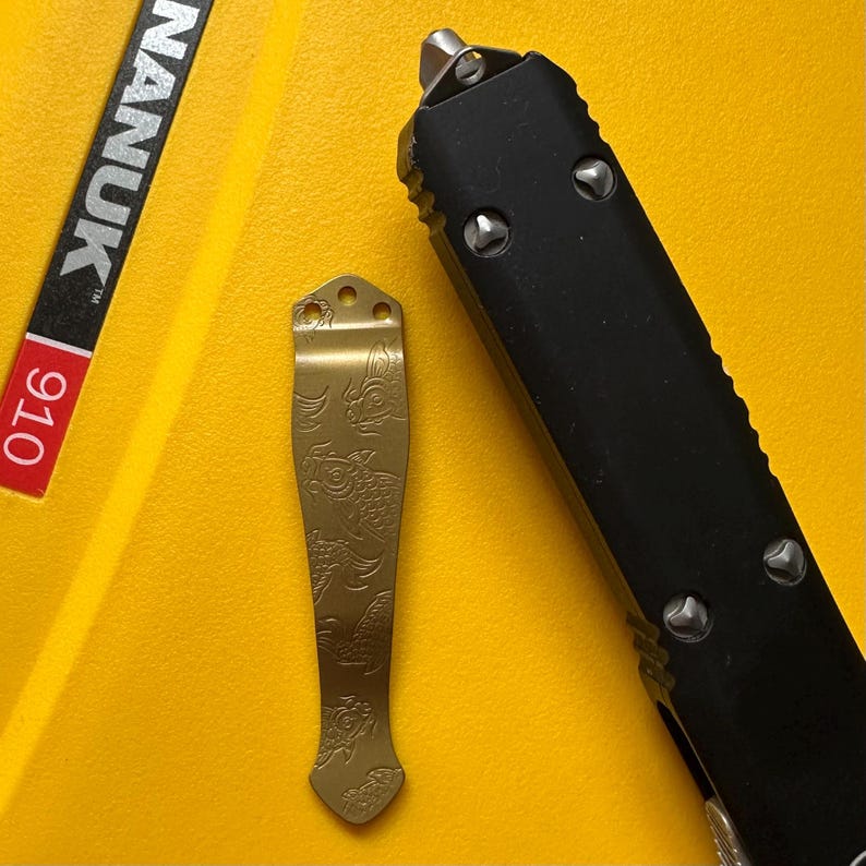 May include: A gold-colored metal pocket clip featuring an engraved koi fish pattern, positioned next to a black handle of an out-the-front knife. The clip has three small circular holes at the top for attachment and a tapered, flared design.