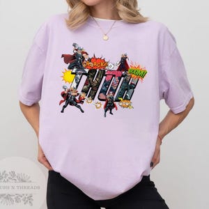 May include: A light purple t-shirt featuring a comic book style graphic of the character Thor. The graphic includes the word "THOR" in large letters, along with action bubbles and illustrations of Thor wielding his hammer.