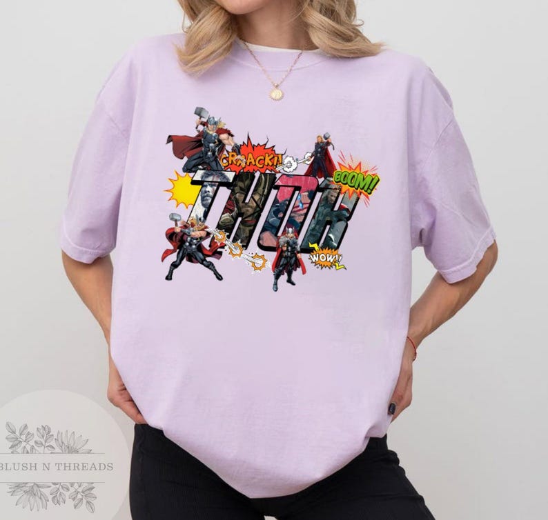 May include: A light purple t-shirt featuring a comic book style graphic of the character Thor. The graphic includes the word "THOR" in large letters, along with action bubbles and illustrations of Thor wielding his hammer.