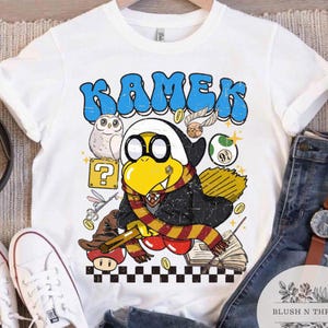 以下が含まれることがあります： 白いTシャツ。青い泡文字で「KAMEK」という文字と、スカーフとメガネをかけた漫画のキャラクターが描かれています。デザインにはフクロウ、疑問符、その他の漫画の要素が含まれています。