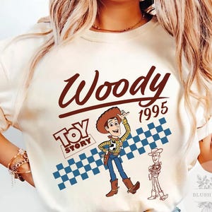 May include: Cream-colored t-shirt featuring a graphic of Woody from Toy Story. The design includes the word "Woody" in a brown script font, the year "1995", and the Toy Story logo. A blue and white checkered pattern is behind the Woody graphic.