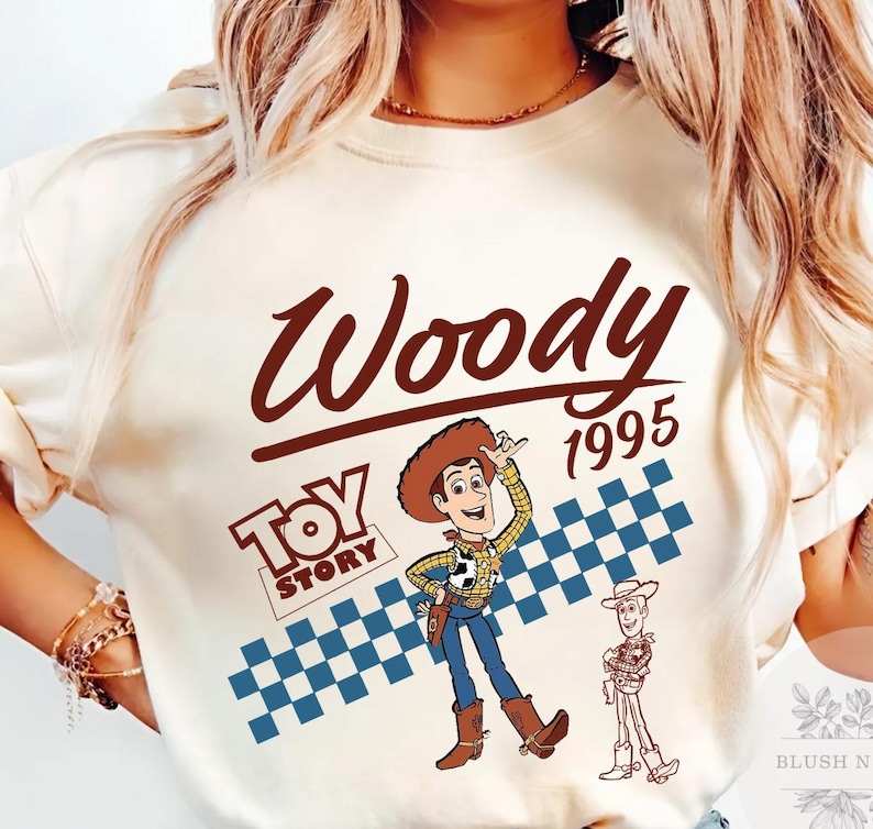 May include: Cream-colored t-shirt featuring a graphic of Woody from Toy Story. The design includes the word "Woody" in a brown script font, the year "1995", and the Toy Story logo. A blue and white checkered pattern is behind the Woody graphic.