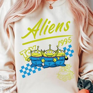 May include: Cream-colored sweatshirt featuring the text "Aliens 1995" in green and a cartoon graphic of three green aliens in blue suits. The design includes a blue and white checkered pattern and the word "Toy".