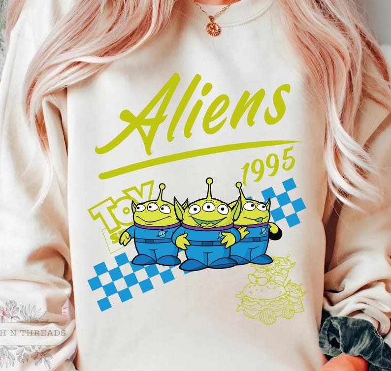 May include: Cream-colored sweatshirt featuring the text "Aliens 1995" in green and a cartoon graphic of three green aliens in blue suits. The design includes a blue and white checkered pattern and the word "Toy".