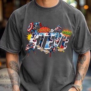May include: Dark gray t-shirt featuring a graphic of Captain America with the name "STEVE" in large, stylized letters. Comic book sound effects like "CRAACK!" and "BOOM!" are also included in the design.