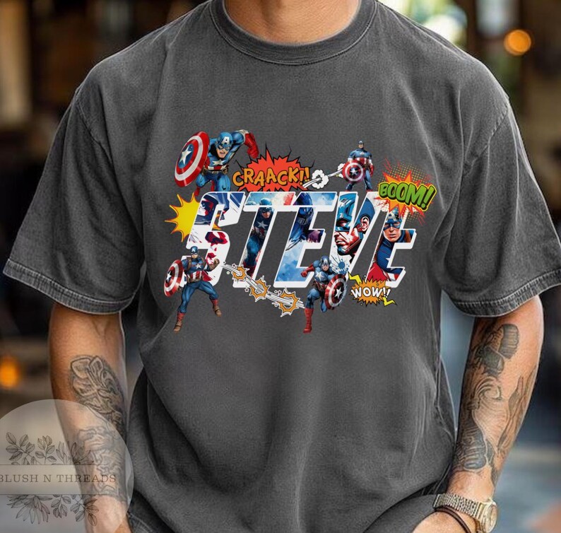 May include: Dark gray t-shirt featuring a graphic of Captain America with the name "STEVE" in large, stylized letters. Comic book sound effects like "CRAACK!" and "BOOM!" are also included in the design.