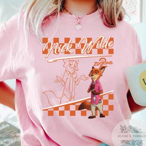May include: Pink t-shirt featuring a cartoon image of Nick Wilde from Zootopia. The design includes the text "Nick Wilde" in an orange script font, a checkered pattern, and the Disney Zootopia logo. The shirt is a light pink color.