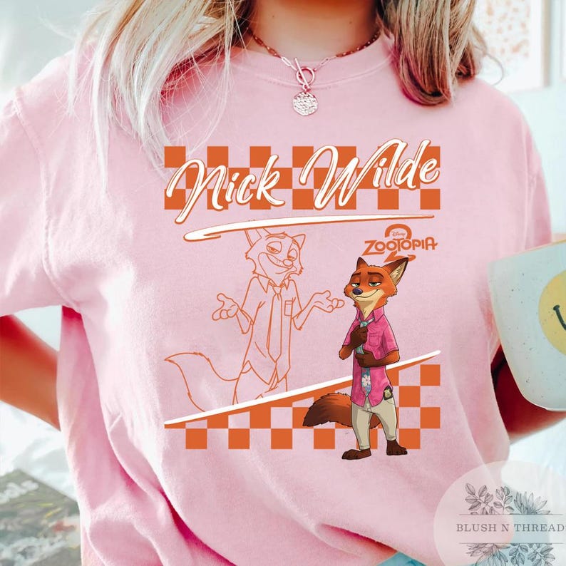 May include: Pink t-shirt featuring a cartoon image of Nick Wilde from Zootopia. The design includes the text "Nick Wilde" in an orange script font, a checkered pattern, and the Disney Zootopia logo. The shirt is a light pink color.