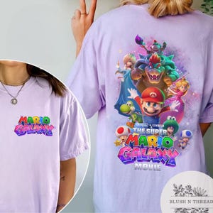 Puede incluir: Una camiseta lila con el texto "MARIO GALAXY" en una fuente 3D colorida en la parte delantera. La parte trasera de la camiseta muestra un gr&aacute;fico de personajes de "The Super Mario Bros. Movie".