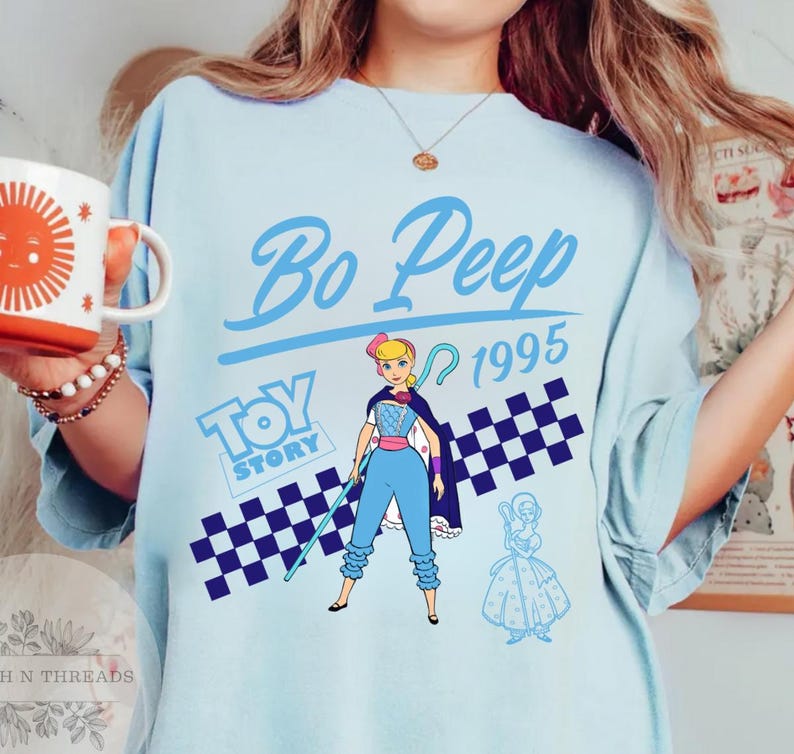 May include: Light blue t-shirt featuring a graphic of Bo Peep from Toy Story, with the text "Bo Peep 1995" in a stylized font. The design includes a checkered pattern and the "Toy Story" logo. A mug with a sun design is visible.