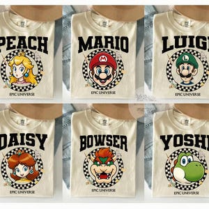 May include: Cream-colored t-shirts featuring characters from the Super Mario video game series. Each shirt displays a character's name in bold black letters above a circular graphic with the character's image and the text "EPIC UNIVERSE".