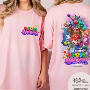 Puede incluir: Camiseta rosa claro con el texto "MARIO GALAXY" en letras coloridas en la parte delantera. La parte trasera de la camiseta muestra un gr&aacute;fico vibrante de Mario y otros personajes de la pel&iacute;cula Super Mario Galaxy.