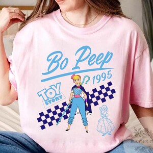 May include: Pink t-shirt featuring a graphic of Bo Peep from Toy Story, with the text "Bo Peep 1995" in blue. The design includes a checkered pattern and the Toy Story logo. The shirt is a light pink color.