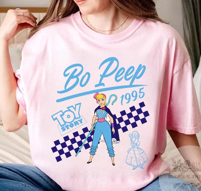 May include: Pink t-shirt featuring a graphic of Bo Peep from Toy Story, with the text "Bo Peep 1995" in blue. The design includes a checkered pattern and the Toy Story logo. The shirt is a light pink color.