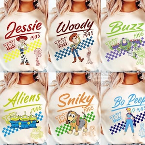 May include: Cream-colored long-sleeve shirts featuring various Toy Story characters: Jessie, Woody, Buzz, Aliens, Slinky, and Bo Peep. Each shirt has the character's name, the year 1995, and the Toy Story logo. The designs include colorful illustrations.