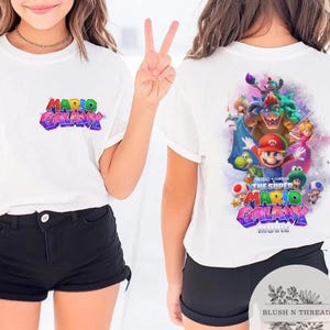 Puede incluir: Camiseta blanca con el texto "MARIO GALAXY" en letras coloridas y estilizadas en la parte delantera. La parte trasera de la camiseta muestra un gr&aacute;fico de Mario y otros personajes de la pel&iacute;cula Super Mario Galaxy.