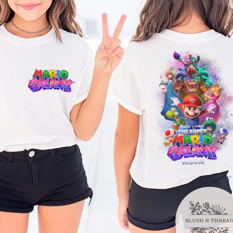 Puede incluir: Camiseta blanca con el texto "MARIO GALAXY" en letras coloridas y estilizadas en la parte delantera. La parte trasera de la camiseta muestra un gr&aacute;fico de Mario y otros personajes de la pel&iacute;cula Super Mario Galaxy.