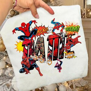 May include: White t-shirt featuring the word "DAD" in a bold, comic book style with images of Spider-Man. The design includes action bubbles with words like "ACK!" and "BOOM!" in vibrant colors.