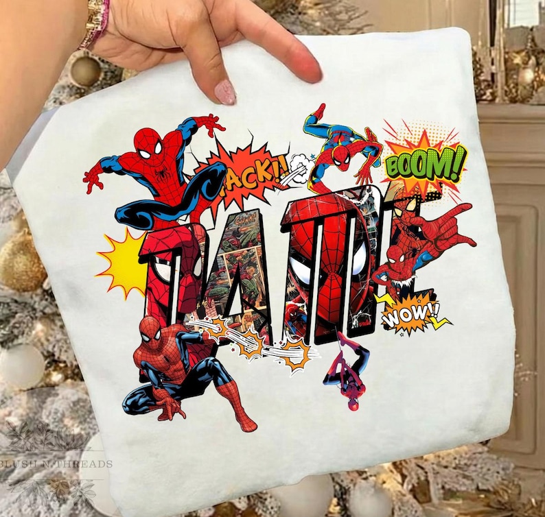 May include: White t-shirt featuring the word "DAD" in a bold, comic book style with images of Spider-Man. The design includes action bubbles with words like "ACK!" and "BOOM!" in vibrant colors.