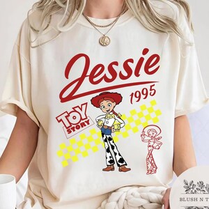 May include: Cream-colored t-shirt featuring a graphic of Jessie from Toy Story. The design includes the name "Jessie" in red script, the year "1995", and the Toy Story logo. A cartoon image of Jessie is also present.