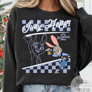 May include: Dark gray sweatshirt featuring Judy Hopps from Zootopia. The design includes a cartoon image of Judy Hopps in a police uniform, a checkered pattern, and the text "Judy Hopps" in white script. The Zootopia logo is also visible.