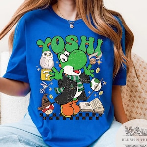 以下が含まれることがあります： ロイヤルブルーのTシャツ。緑色の恐竜キャラクター、ヨッシーのグラフィックが、魔法使いをテーマにしたデザインで描かれています。デザインにはフクロウ、キノコ、緑色の「ヨッシー」の文字、その他の漫画の要素が含まれています。