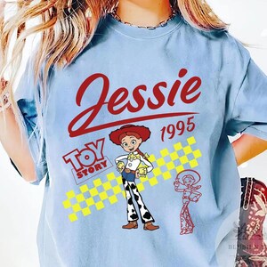 May include: Light blue t-shirt featuring the name "Jessie" in red script above the year "1995". The shirt includes a "Toy Story" logo and a cartoon image of Jessie, the cowgirl, along with a smaller outline of her. A yellow and white checkered pattern is also present.