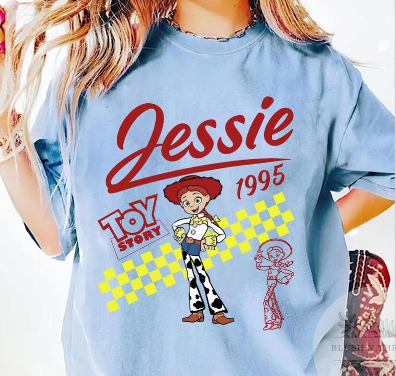 May include: Light blue t-shirt featuring the name "Jessie" in red script above the year "1995". The shirt includes a "Toy Story" logo and a cartoon image of Jessie, the cowgirl, along with a smaller outline of her. A yellow and white checkered pattern is also present.