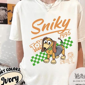 May include: Ivory-colored t-shirt featuring a cartoon image of Slinky Dog from Toy Story. The design includes the word "Sniky" in orange script, the year "1995", and the Toy Story logo. A green and white checkered pattern is behind the dog.
