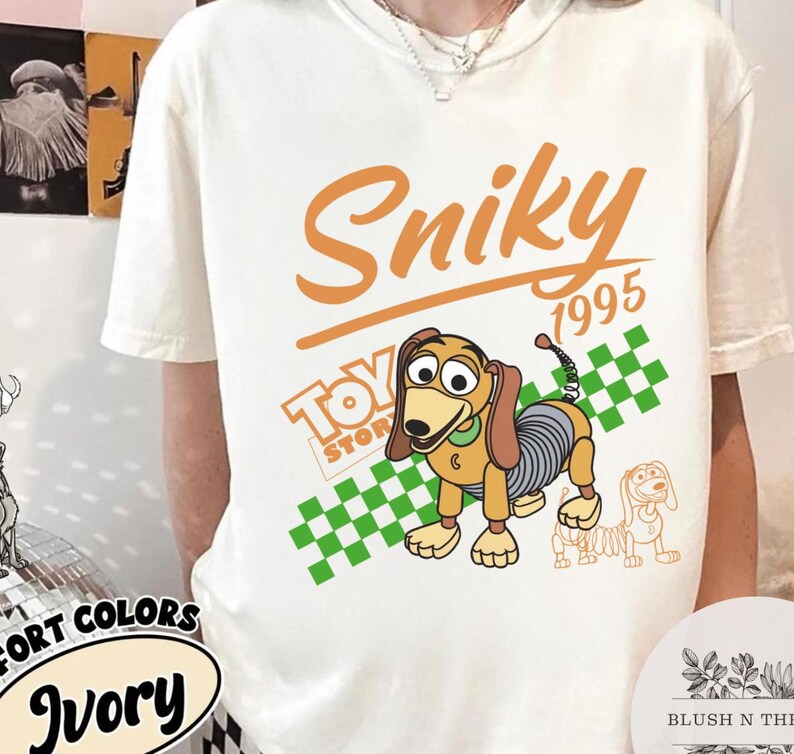 May include: Ivory-colored t-shirt featuring a cartoon image of Slinky Dog from Toy Story. The design includes the word "Sniky" in orange script, the year "1995", and the Toy Story logo. A green and white checkered pattern is behind the dog.