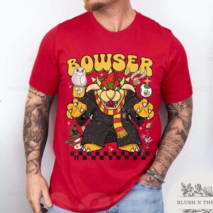 以下が含まれることがあります： 赤色のTシャツ。黄色で「BOWSER」の文字と、フクロウ、クエスチョンマーク、その他の漫画の要素が描かれた、Bowserのグラフィックが特徴です。デザインは、使い古されたようなヴィンテージ感があります。