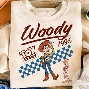 May include: Cream-colored sweatshirt featuring a graphic of Woody from Toy Story. The design includes the word "Woody" in a brown script, the year "1995", and the "Toy Story" logo. A blue and white checkered pattern is also present.