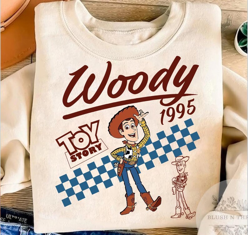 May include: Cream-colored sweatshirt featuring a graphic of Woody from Toy Story. The design includes the word "Woody" in a brown script, the year "1995", and the "Toy Story" logo. A blue and white checkered pattern is also present.