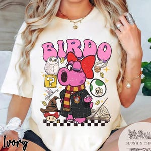 以下が含まれることがあります： ピンク色のバードキャラクターが赤いリボン、スカーフ、黒いローブを着たアイボリー色のTシャツ。デザインにはフクロウ、疑問符、その他の気まぐれな要素が含まれています。「BIRDO」という言葉がキャラクターの上に印刷されています。