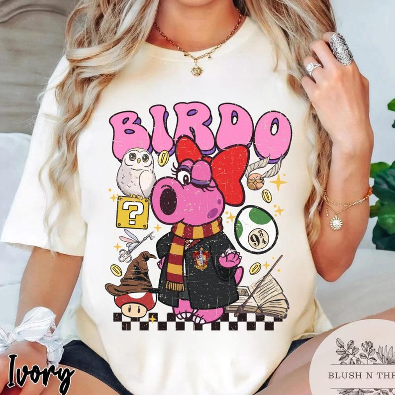 以下が含まれることがあります： ピンク色のバードキャラクターが赤いリボン、スカーフ、黒いローブを着たアイボリー色のTシャツ。デザインにはフクロウ、疑問符、その他の気まぐれな要素が含まれています。「BIRDO」という言葉がキャラクターの上に印刷されています。